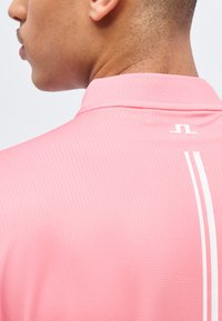 Κοντινό πλάνο στον λαιμό και τον ώμο ενός ατόμου που φοράει ροζ πουκάμισο με υφή, με ένα μικρό λευκό λογότυπο και δύο κάθετες ρίγες στην πλάτη.