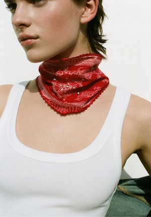 Zbliżenie osoby noszącej biały top i czerwony wzorzysty bandana szalik wokół szyi na jednolitym tle.