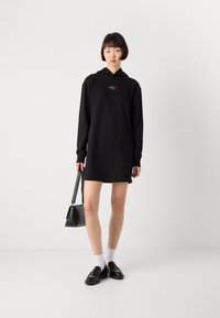 Čierna oversized mikinová šaty s kapucňou a dlhými rukávmi, predným logom, hladkým materiálom a bočnými vreckami. V kombinácii s čiernymi topánkami a taškou.