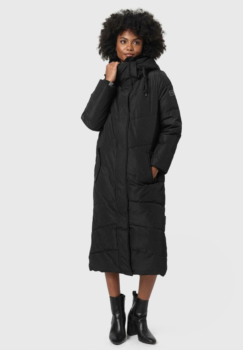 Long manteau noir rembourré avec un col haut, fermeture éclair, poches latérales et détails cousus, porté avec des bottines noires.
