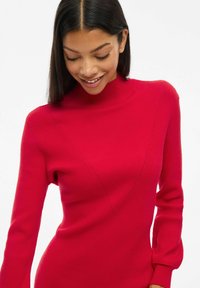 Robe en tricot rouge à mailles côtelées, à manches longues, avec un col haut et un design en V sur le devant, présentant une texture lisse et une silhouette ajustée.
