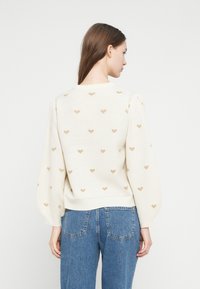 Κρεμ πουλόβερ με χρυσά σχέδια καρδιάς, με φουσκωτά μανίκια και ριμπ τελείωμα. Φορεμένο με τζιν παντελόνια ψηλόμεσα μπλε, που δείχνουν casual εφαρμογή.