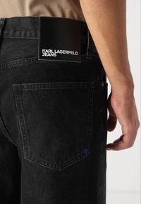 Black denim jeans with a smooth texture, featuring a black leather patch labeled "KARL LAGERFELD JEANS" on the back. Accented with a small blue "K."