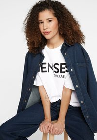 Denim jacket over white T-shirt with "SENSES" print. Dark denim pants. Curly hair, light skin. Simple background without distractions.