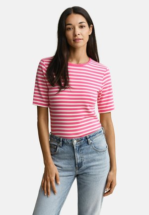 Frau mit langen dunklen Haaren, die ein kurzärmeliges, rosa-weiß gestreiftes Shirt und hellblaue, hoch taillierte Jeans trägt, vor einem weißen Hintergrund.