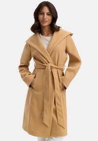 Beige wikkeljas met een grote capuchon, strikband en zijzakken; heeft een zachte textuur en een reliëf merkdetail op de mouw.