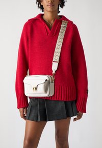 Maglione rosso lavorato a maglia con scollo a V, abbinato a una borsa a tracolla bianca in pelle con chiusura dorata e tracolla decorata. Gonna a scacchi nera.