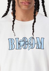 Hvit bomull t-skjorte med ordet "BLOOM" i fet svart og lyseblå skrift og med et blomsterdesign.