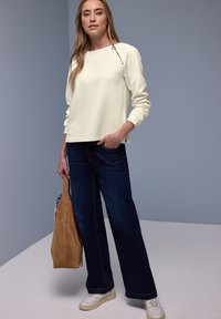 Jeune femme portant un haut blanc texturé à manches longues avec une fermeture éclair sur l'épaule, un jean large foncé, des baskets blanches, tenant un sac en daim marron.