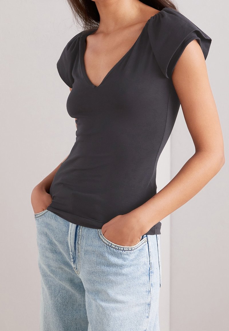 Femme portant un haut noir cintré à col en V avec des manches courtes bouffantes et un jean en denim bleu clair, les mains dans les poches.