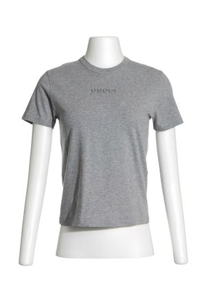 Camiseta gris de manga corta con cuello redondo y logotipo "Gucci" sutilmente en relieve en el pecho, mostrada en un torso de maniquí blanco.
