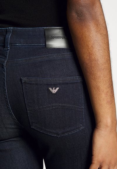 Tummansiniset farkut mustalla nahkavyöllä, jossa on kohokuvioituna "EMPORIO ARMANI". Huomattava hopeinen logo takataskussa.