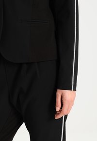 Černá sako s elegantním designem, obsahující jedno náprsní kapsu a stříbrný pruh podél rukávů. Viditelná je hladká textura látky.