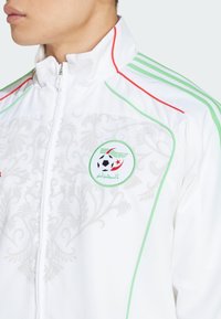 Vit sportjacka med gröna och röda detaljer, med ett blommigt mönster, dragkedjestängning, hög krage och en cirkulär logotyp med en fotboll.