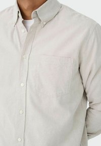 Lys beige skjorte med knapper og krave, brystlomme og hvide knapper. Lavet af et blødt stof med en glat overflade.