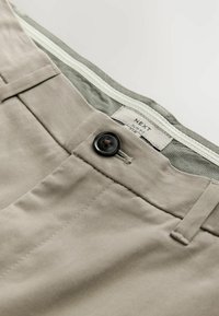 Világos bézs színű, slim fit nadrág sima textúrával, fekete gombbal, cipzárral és a derékrész belsejébe varrott címkével.