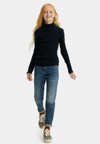 Jūras spēku zaļa velūra torte, zili džinsi un leopardu raksta kedas. Apģērbs ir ar pieguļošu dizainu un klasisku džinsu stilu ar sarullētām piedurknēm.