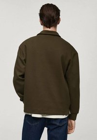 Felpa verde oliva scuro con vestibilità comoda, dotata di colletto e cuciture a vista. Indossata sopra una camicia bianca e abbinata a jeans blu.