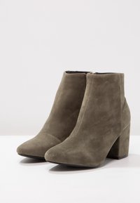 Botas de tobillo de ante en verde oliva con punta afilada, tacón alto en bloque y cierre de cremallera en la parte trasera. Textura suave y costuras mínimas.