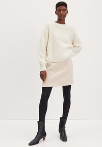 Cremfarbener gestrickter Pullover mit lockerem Schnitt, kombiniert mit einem hellbeigen Minirock, schwarzen Strumpfhosen und spitzen Ankle Boots.