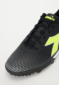 Botas de fútbol negras con parte superior texturizada, logo de flecha verde y patrón de puntos plateados en la parte delantera. Cuenta con cordones negros y suela con tacos.