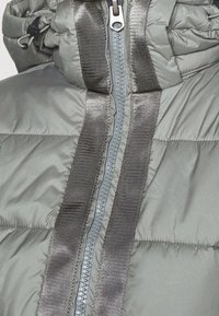 Chladící bunda v šedé barvě s prošívanou texturou. Disponuje centrálním zipem a dvěma širokými, lesklými černými pásy vpředu.