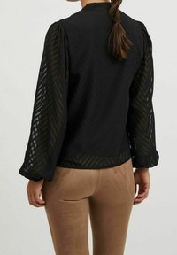 Blusa nera con motivo a chevron testurizzato, maniche lunghe trasparenti e collo alto. Indossata con pantaloni aderenti beige chiaro.