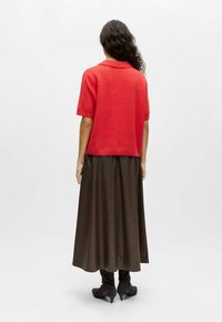 Pull rouge à manches courtes, col rond, associé à une jupe longue marron, texture lisse, et bottes noires en daim à talons hauts.