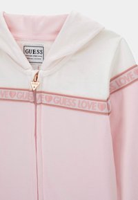 Felpa con cappuccio rosa chiaro con cerniera, con cappuccio bianco. Presenta una banda logo "GUESS LOVE" in un rosa a contrasto e una cerniera color oro rosa. Tessuto morbido.