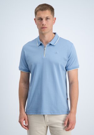 Uomo che indossa una polo azzurro chiaro a maniche corte con colletto con cerniera e pantaloni beige, in piedi davanti a uno sfondo chiaro uniforme.
