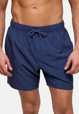 Hombre vistiendo pantalones cortos de baño azul marino con pequeños patrones de anclas y un cordón atado en la cintura, recortados a la mitad del muslo.