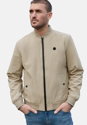 Mann mit kurzem Haar, der eine beige Bomberjacke mit Reißverschluss, schwarzen Knöpfen und blauer Jeans trägt, die Hände in den Taschen, schaut zur Seite.