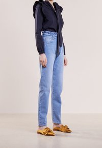 Blusa blu scuro con scollatura arricciata, jeans azzurri a vita alta e scarpe slip-on dorate con dettaglio a nodo. Texture complessive morbide.