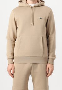 Sweat à capuche beige avec poche avant et cordons, associé à un short beige assorti, arborant un petit logo crocodile vert sur la poitrine.