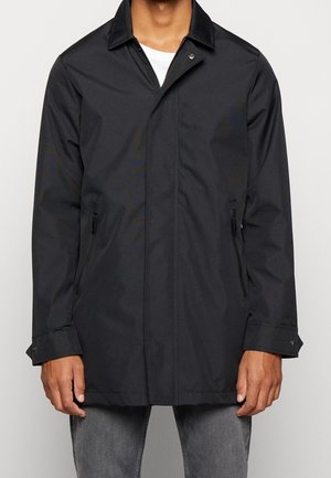 Homme portant une veste noire zippée avec col et poches latérales zippées, associée à un jean gris et une chemise blanche en dessous.
