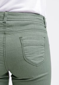 Vihreät farkut, joissa on takatasku hienovaraisilla ompeluyksityiskohdilla ja keskikorkea vyötärö. Silkkinen tekstuuri ja muotoiltu malli ovat havaittavissa.