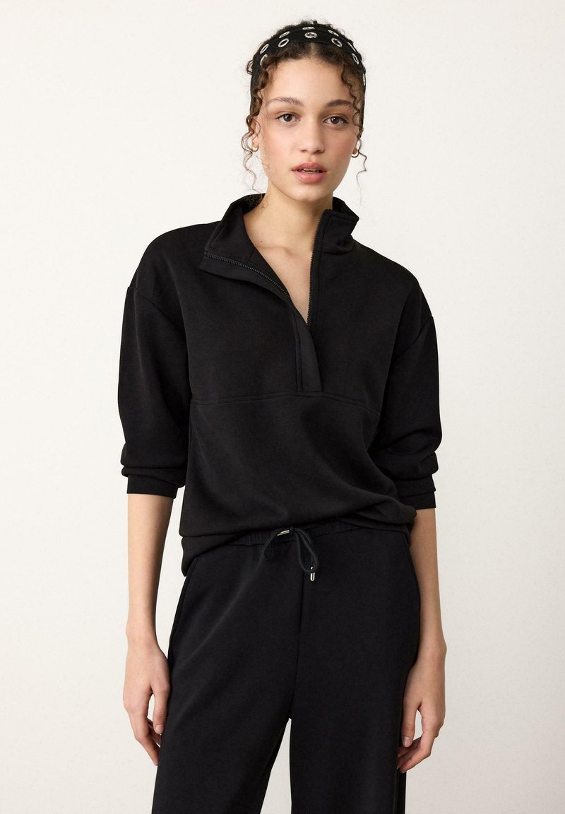 Schwarzer Pullover mit Reißverschluss aus weichem Stoff und hohem Kragen, mit weiten Ärmeln und lockerer Passform, kombiniert mit passenden schwarzen Jogginghosen.