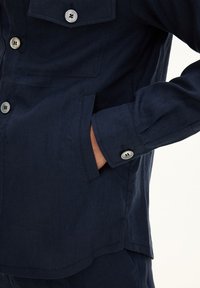 Navy blue linen shirt with a front pocket, buttoned cuffs, and silver buttons. Features a relaxed fit and smooth texture.