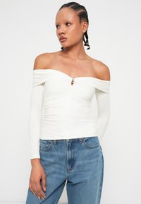 Top blanco con los hombros al descubierto y mangas largas, con un escote central en forma de llave y detalle metálico. Combinado con jeans azul de tiro alto.