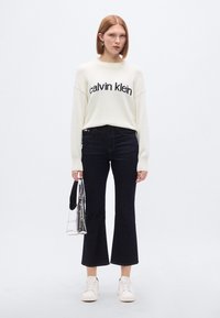 Ανοιχτό κρεμ πουλόβερ με λογότυπο "Calvin Klein", σκούρο καμπάνα τζιν, λευκά αθλητικά παπούτσια και ασημένια μεταλλική τσάντα. Λεία υφή και casual στυλ.