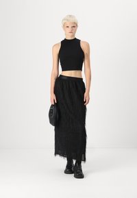 Black sleeveless crop top, textured black maxi skirt with fringe detail, black footwear, and a small black handbag. Smooth floor backdrop.