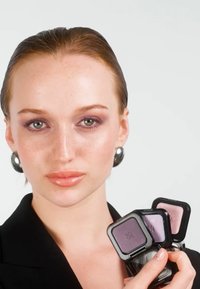 Femme aux cheveux lissés en arrière et portant des boucles d'oreilles argentées tient près de son visage trois teintes de fard à paupières violet, vêtue d'un haut noir.