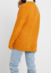 Maglione arancione di grandi dimensioni in maglia con finitura testurizzata, polsini e orlo a coste, abbinato a jeans azzurri chiari. Vista posteriore che mostra il design.