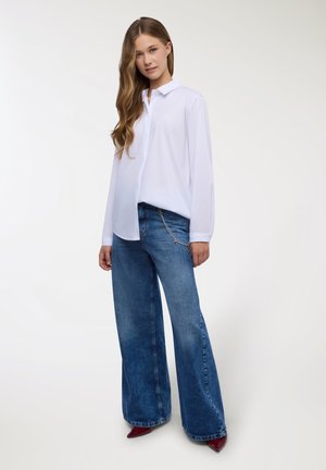 Camicia bianca a maniche lunghe con colletto abbottonato abbinata a jeans di jeans blu a gamba larga con accessorio a catena sull'anca.