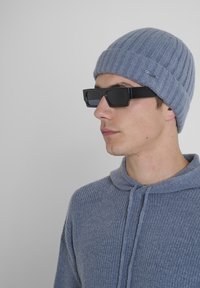 Svijetloplava rebrasta pletena kapa, u paru s pravokutnim crnim naočalama. Model nosi sivi strukturirani pulover s kapuljačom na vezanje.