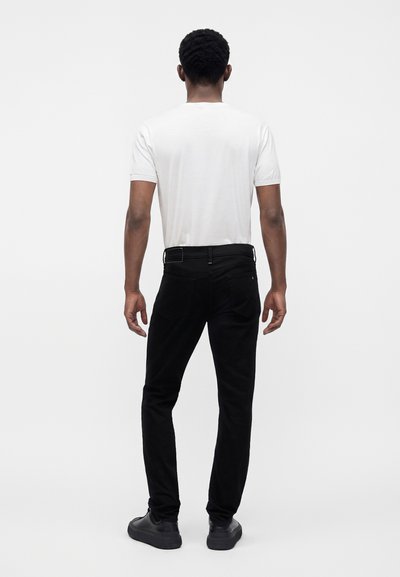 Homme debout de dos, portant un t-shirt blanc uni à manches courtes, un pantalon noir et des chaussures noires sur un fond clair uni.