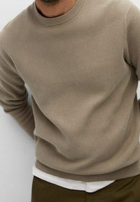 Beżowy sweter z prążkowanej tkaniny, z okrągłym dekoltem i długimi rękawami, nałożony na białą koszulę. Miękka tekstura, dopasowany krój i czyste linie.