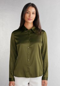 Blusa in satin di un verde oliva scuro, con maniche lunghe, chiusura frontale a bottoni e colletto appuntito. Texture liscia con orlo curvo e bottoni coordinati.