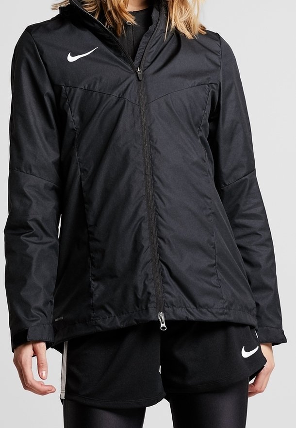 Personne portant une veste zippée noire Nike avec le logo sur la poitrine, associée à un short Nike noir et des leggings noirs.