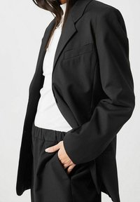 Zwarte blazer van gladde stof, met een enkele borstzak, brede revers en verlengde mouwen, gecombineerd met een witte top en zwarte broek.